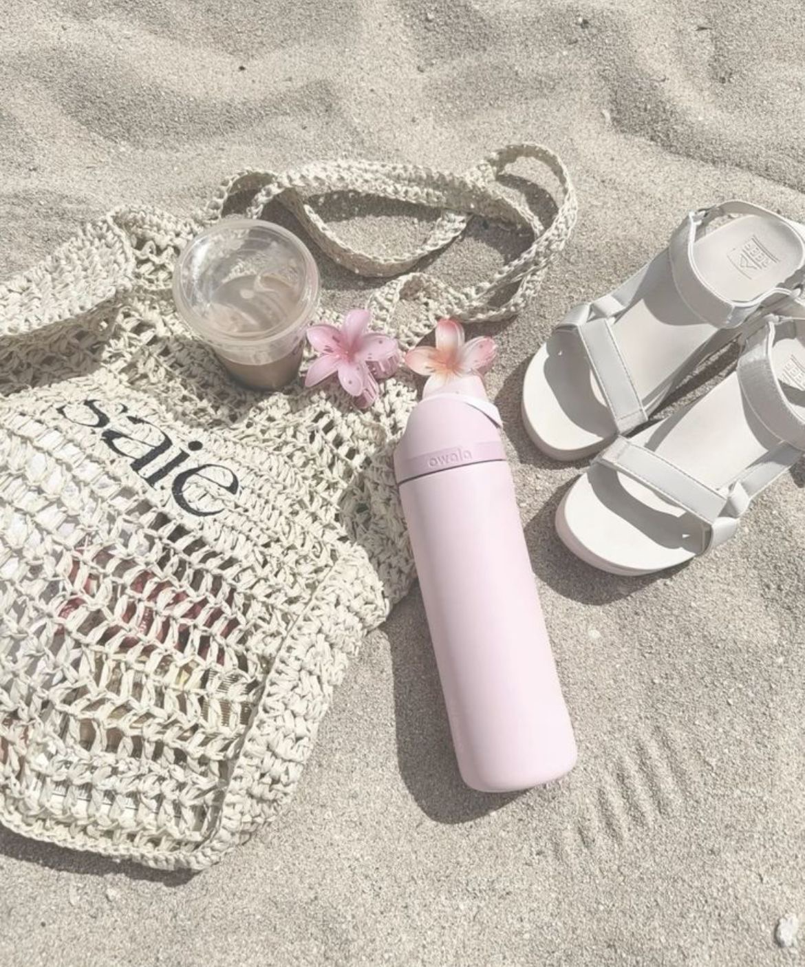 Accessoires de plages déposés sur le sable, comprenant un sac en maille, une gourde, des sandales et des fleurs.