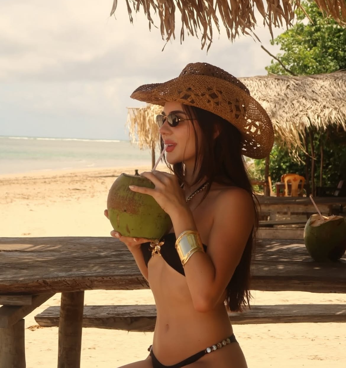 Une modèle de profil à la plage portant un bikini deux-pièce noir, un chapeau et des lunettes de soleil.